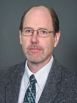 A person in glasses looks at the camera while wearing a gray tweed jacket and teal patterned tie in front of a gray backdrop