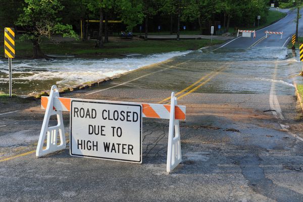 A road closed to due to high water sign attached to a sawhorse sits in front of a flooded roadway.