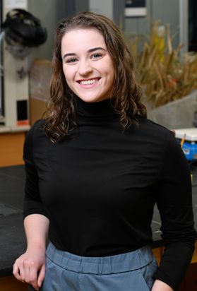 Headshot of WVU student Teagan Kusniar. She is pictured standing in a lab wearing a black turtleneck and blue-gray pants. She has long, curly brown hair. 