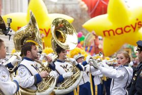 More than a dozen band members are shown on the streets of New York in front of Macy's balloons. Those in the front are playing tubas. A conductor wears a white uniform on the right side of the frame.