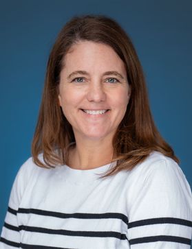 Headshot of Lesley Cottrell. She is wearing a blue and white striped sweater. She has long brown hair and is pictured in front of a blue background. 