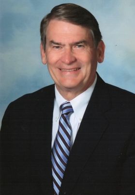Headshot of Glenn Whisler. He is pictured against a blue background and is wearing a dark colored suit with a white dress shirt and a blue striped tie. He has short, dark hair. 