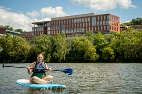A woman sits on top of a paddle board holding her paddle with a red bricked building on the WVU Campus behind her. 