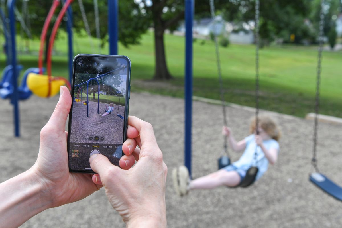 A pair of hands holds a phone being used to take a photo of a child with blond hair swinging on a playground. 