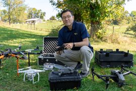 WVU professor Yong Lak-Park is shown here surrounded by drones that he uses in his research. He is crouched near to the ground and is holding one of the controllers in his hands. 