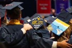 A photograph taken of the back of a group of recent WVU graduates arm in arm during the Commencement ceremony. Two of the mortar boards in the frame are creatively decorated. One with the words: The best view comes after the hardest climb." 