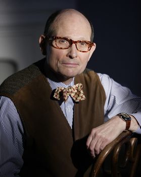 A man with brown rectangular glasses wearing a striped blue and white button up shirt with a brown vest jacket and a brown plaid bow tie