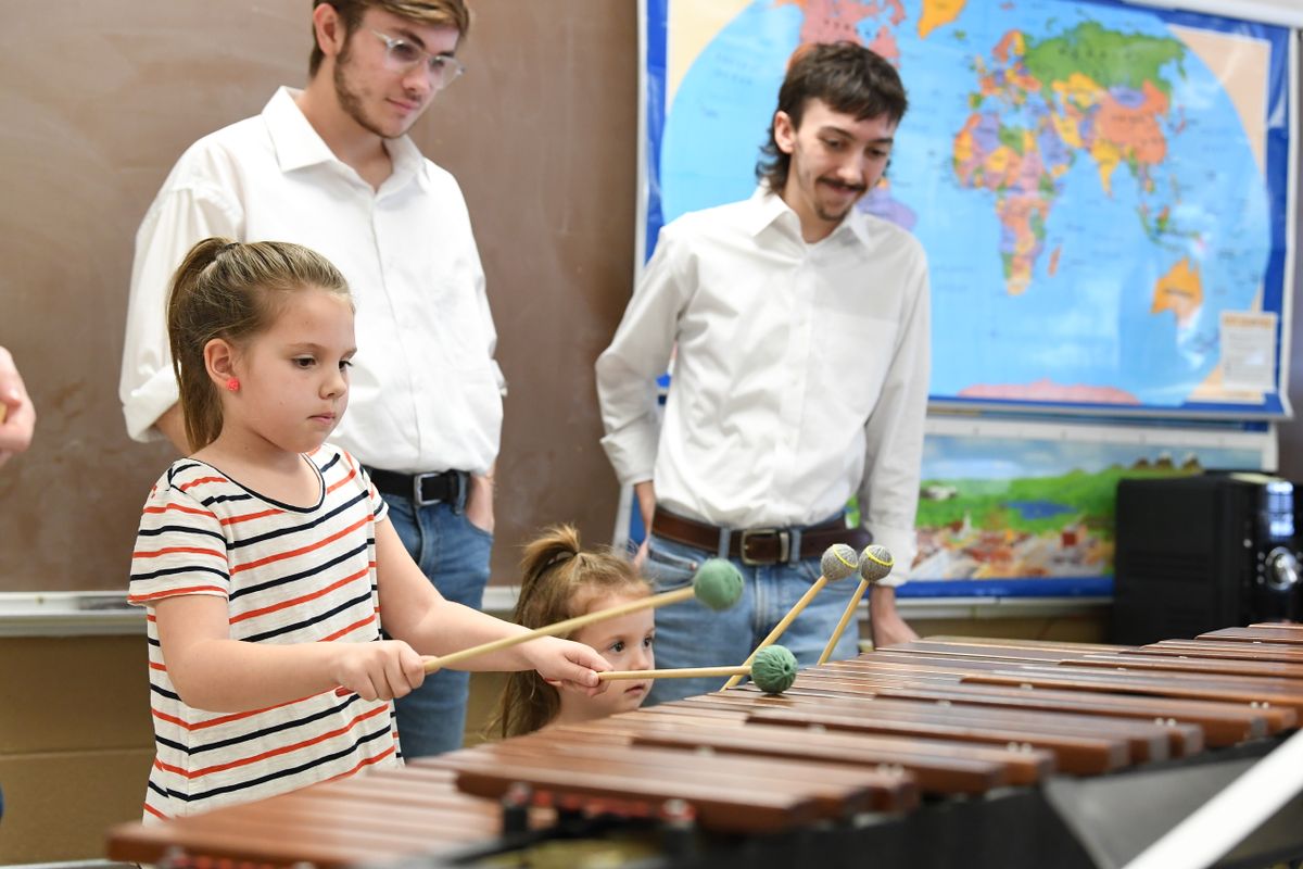 A picture of two young girls, one with a pony tail wearing a striped shirt, and the other barely tall enough to see over the instrument, playing the xylophone. Two older gentlemen are watching as they play. 