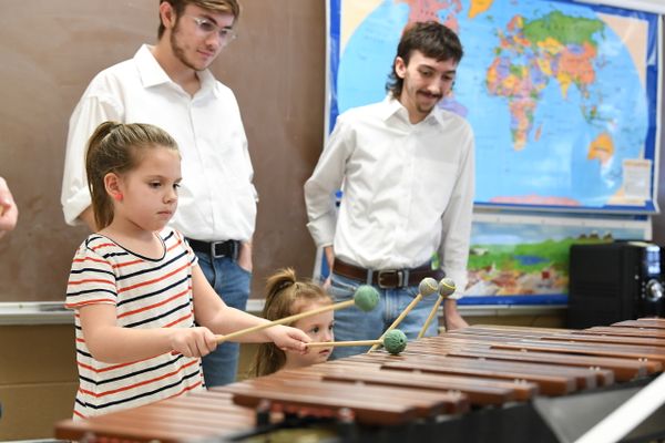 A picture of two young girls, one with a pony tail wearing a striped shirt, and the other barely tall enough to see over the instrument, playing the xylophone. Two older gentlemen are watching as they play. 