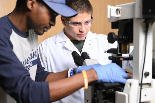 Two male researchers work together in a lab using a microscope. One researcher is wearing a blue and white baseball tee and the other is dressed in a white lab coat and is wearing safety glasses. 