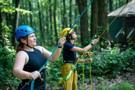 Two women wait at the bottom of a repel line with ropes attached to them. They are each wearing protective helmets. 