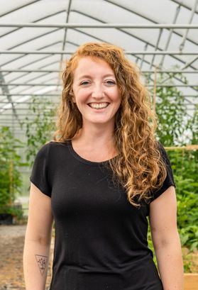 Photograph of scholarship recipient Tiffany Strange. She is pictured in a green house with plants behind her. She is wearing a black T-shirt and has curly, strawberry blonde hair. 
