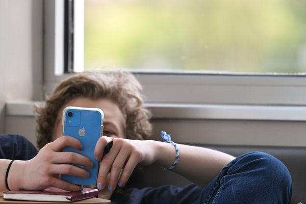 A student slouches in a char with the back of their head barely reaching a window. They are holding a blue iPhone in two hands that is almost entirely hiding their face. 