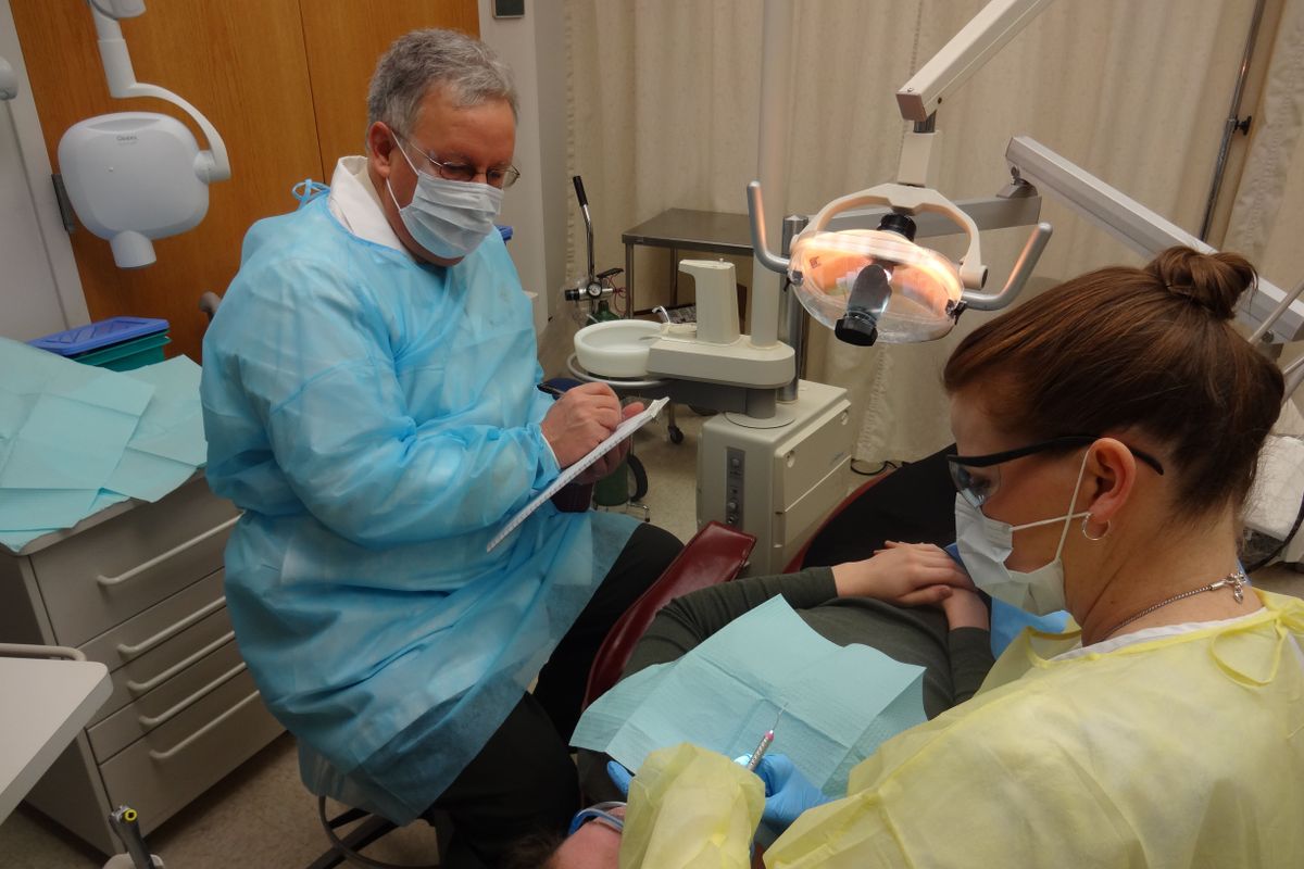 Two people wearing surgical gowns and face masks giving a patient a dental exam.