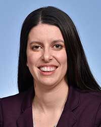 A woman with black hair wearing a plum colored jacket smiles for a headshot