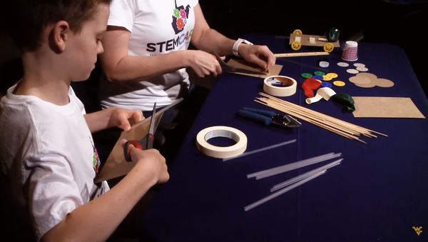 A photograph showing two children completing a STEM activity with pieces of the experiment strewn across a black table. 