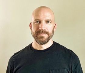 Headshot of William Franko. He is wearing a black tshirt and is standing in front of a tan background. He is bald and had a full brown beard. 