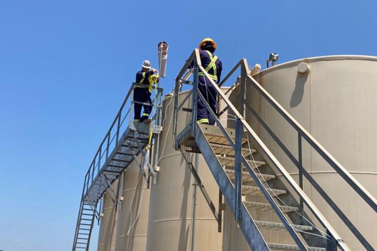 This picture is shot from the ground, looking up at a beige tank with silver stairs to access the top. Two people are at the top in blue work uniforms with bright yellow suspenders and hard hats.