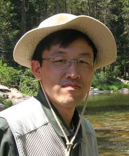A man in thin glasses, a green shirt, a gray fishing vest and a beige hat with chin strap stands in front of a stream