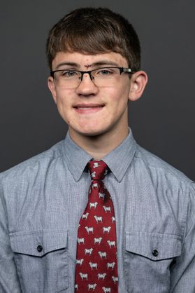 A person wearings a gray button-up shirt, red patterned tie and glasses in front of a gray backdrop.