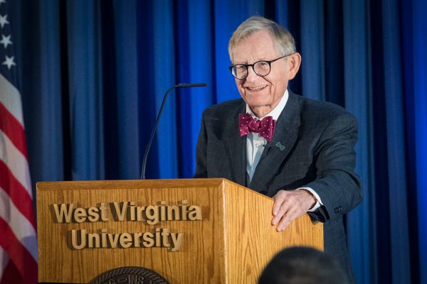 WVU President Gordon Gee stands in front of a navy blue curtain and behind a wooden podium that says 'West Virginia University.' Gee is wearing a dark jacket, red bow tie and black rimmed glasses.