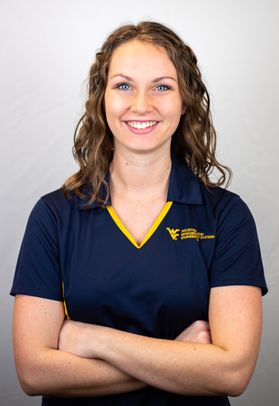 Portrait of Makenzie Dolly, she has shoulder length brown hair and is smiling. She's wearing a WVU collared shirt and stands with her arms crossed.