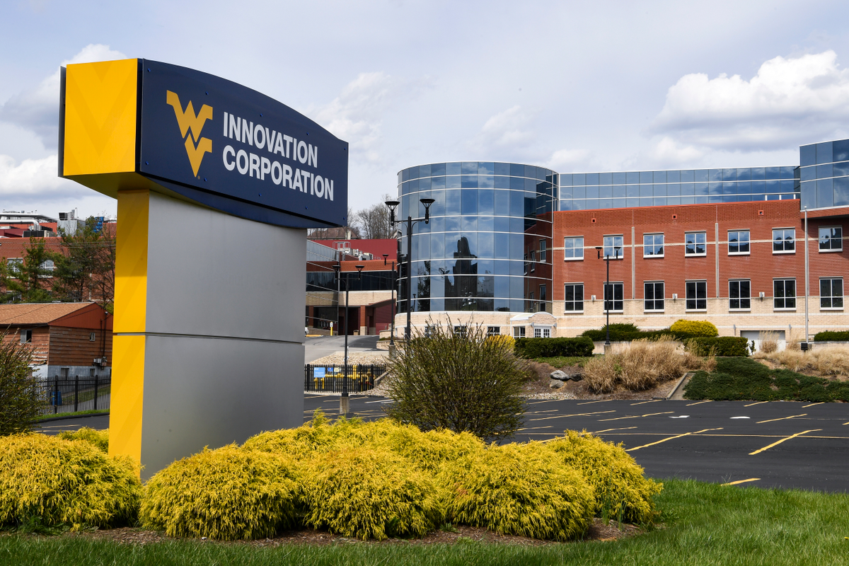 A gray sign is in the front of this shot. It has a Flying WV on it it and the words 'Innovation Corporation.' In the background, a brick building with gray windows is visible. Bushes are around the sign.