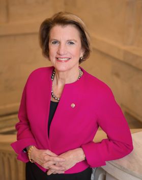 Headshot of U.S. Sen. Shelley Moore Capito. She is pictured inside wearing a magenta jacket over a black dress and with her hands clasped at her waist. She has short blond hair and is smiling. 