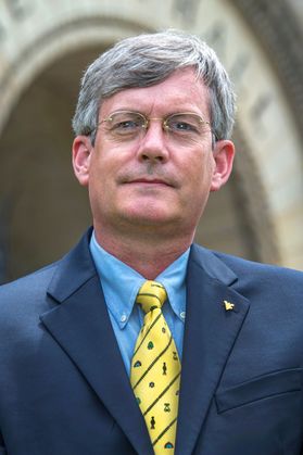 A barely smiling man in dark blue jacket, blue shirt, yellow tie with blue diagonal stripes