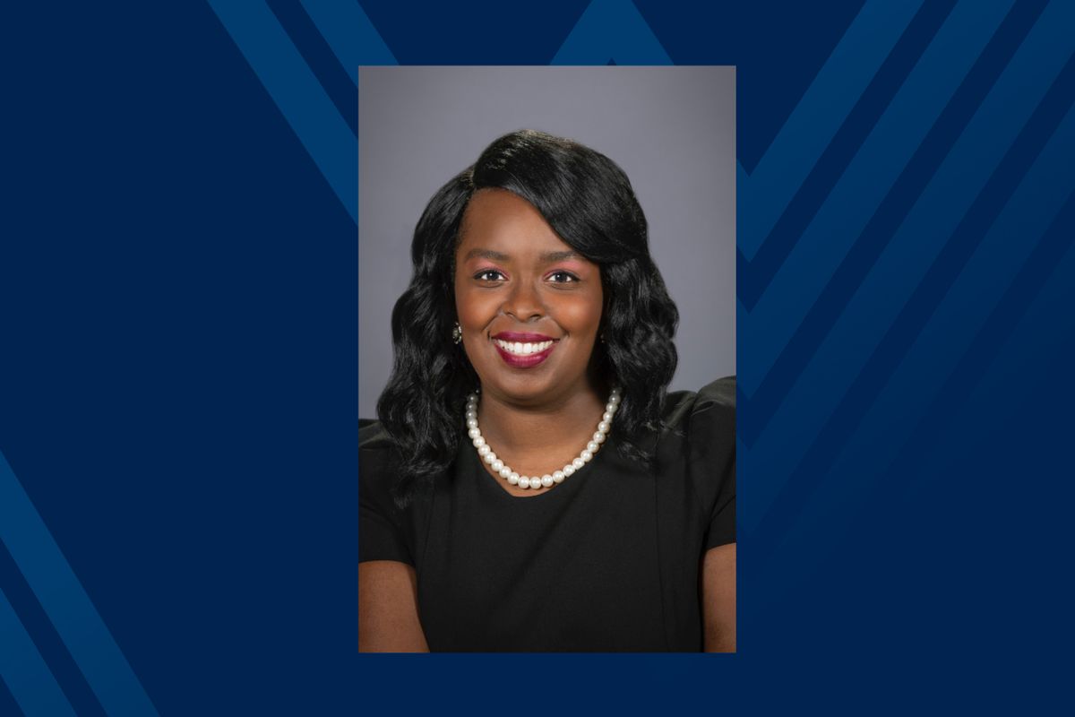 Headshot of smiling Black woman in pearls and black dress on blue background