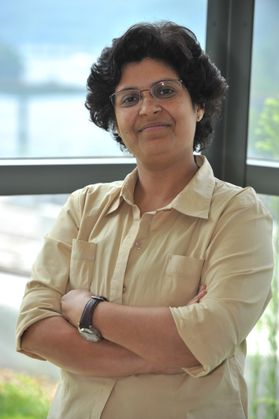 Headshot of Shika Sharma. She is pictured against a glass and is wearing a light colored shirt. She has short, curly dark hair and wearing glasses. 