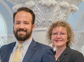 Man with dark hair and beard wearing blue suit and yellow tie standing with blonde woman with curly hair wearing a grey suit jacket 