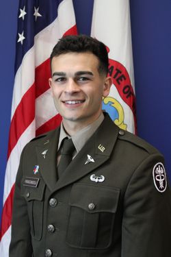 This is a portrait of Lowell Parascandola who has short dark hair and is wearing a green military uniform while standing in front of an American flag.