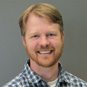 Headshot of Danny Twilley. He is wearing a blue plaid shirt over a white undershirt. He is smiling and has strawberry blond hair with a reddish colored beard. He is pictured against a gray background. 
