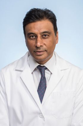 Man with dark black hair poses for a headshot wearing a white button down and a navy blue tie covered by a white lab coat.