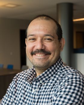 Headshot of WVU professor Bernardo Quiroga. He is pictured inside wearing a dark blue checked shirt and is standing with his arms crossed. He has a brown mustache and very short brown hair. 