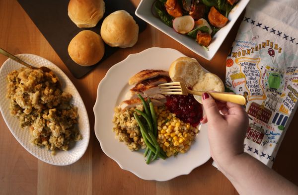 A plate of holiday food is shown I this photograph as is a woman's hand holding a gold-tone fork. On the main white plate are green beans, corn and turkey. Smaller dishes on the table hold stuffing, rolls, and colorful potatoes. 