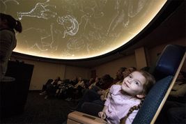 Image of a little girl wearing a long sleeve pink shirt with brown hair looking up at the ceiling in the WVU planetarium. The room is dark and there are many people sitting in chairs 
