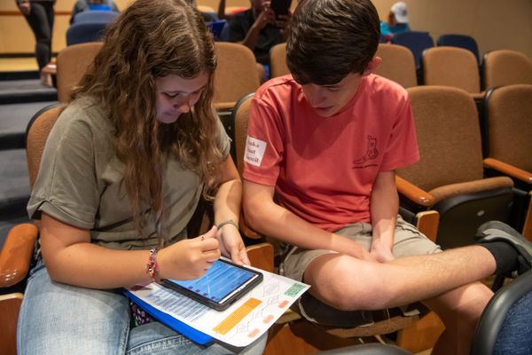 A male student in a red shirt and shorts and a female student in a green shirt and jeans use a tablet to work while in an auditorium. 