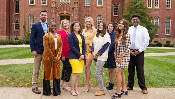 Picture of the nine member of the 2022 WVU Homecoming Court. They are all dressed in business casual clothes and are standing outside in front of Woodburn Hall. 