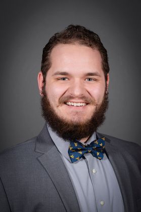 smiling young man with beard wearing gray jacket over light gray shirt and bold and blue bowtie