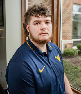 Headshot of WVU Mountaineer mascot candidate Cade Kincaid. He is pictured in front of a window wearing a navy blue WVU-branded golf shirt. He has curly, light brown hair cut in a mullet and a light brown beard. 