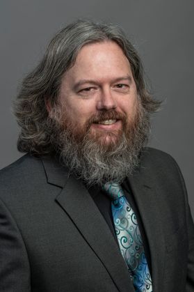 photo of fully bearded man wearing gray jacket, black shirt iridescent green and blue tie