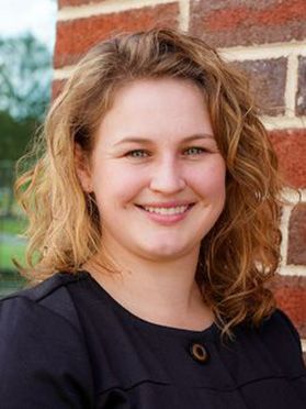 Headshot of WVU professor Samantha Ross. She is standing in front of a red brick wall and there are trees in the background. She is wearing a dark colored top and has dark blonde wavy hair. 