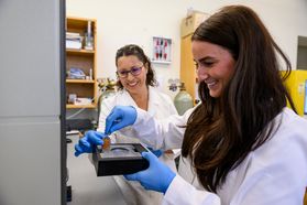 Photograph of two female WVU researchers working together in a lab. Both women have brown hair and are wearing white lab coats and blue surgical gloves. One researcher is holding a piece of lab equipment while the other adds something to it. 
