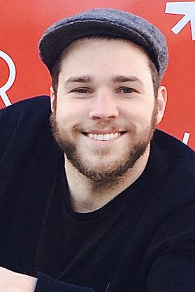 Photograph of Ted Gardener, grandson of WVU donor Margaret Workman. He is pictured in front of a red sign wearing a black T-shirt and wearing a gray paperboy cap. He has a full brown beard.