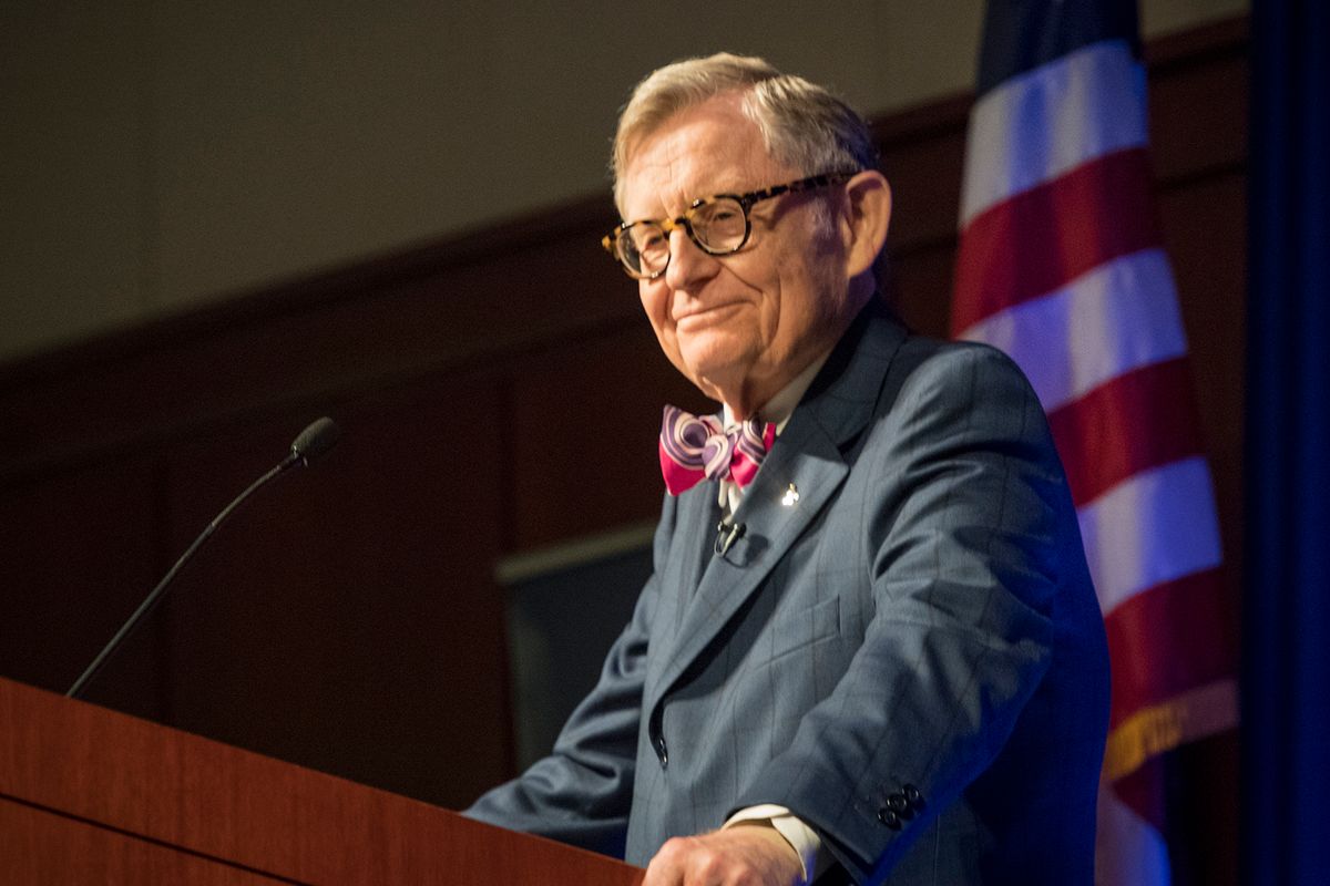 Man in a bow tie and glasses stands behind podium, in front of American flag