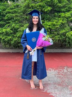 Scholarship recipient Juliana Perdue is pictured here in a blue cap and gown holding a bouquet of flowers wrapped in pink paper and her blue diploma cover. 