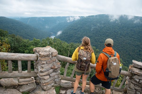 A man and a woman, both wearing casual hiking clothes and backpacks, stand at the overlook in Coopers Rock State Forest looking down into the tree covered landscape of the Cheat River canyon. 