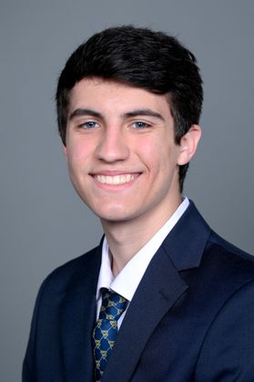 Headshot of WVU Bucklew Scholar Garrett Hutson. He is pictured against a gray background and is wearing a navy blue jacket over a white dress shirt and a blue patterned tie. He has short dark hair. 